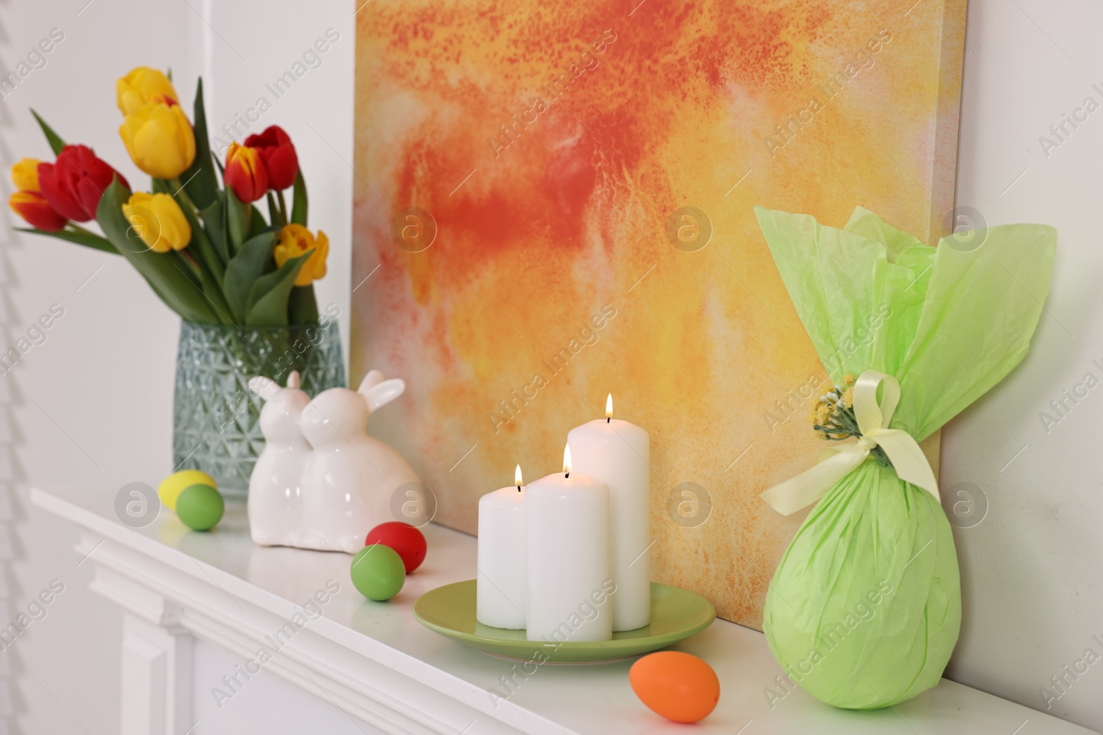 Photo of Easter decorations. Bouquet of tulips in vase, burning candles and bunny figures on fireplace at home