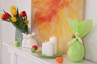 Photo of Easter decorations. Bouquet of tulips in vase, burning candles and bunny figures on fireplace at home