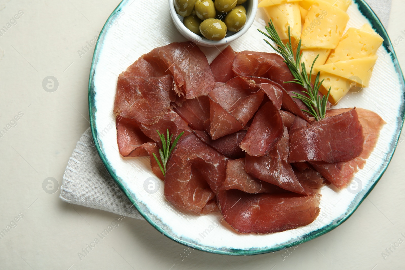 Photo of Delicious bresaola, cheese, olives and rosemary on light textured table, top view