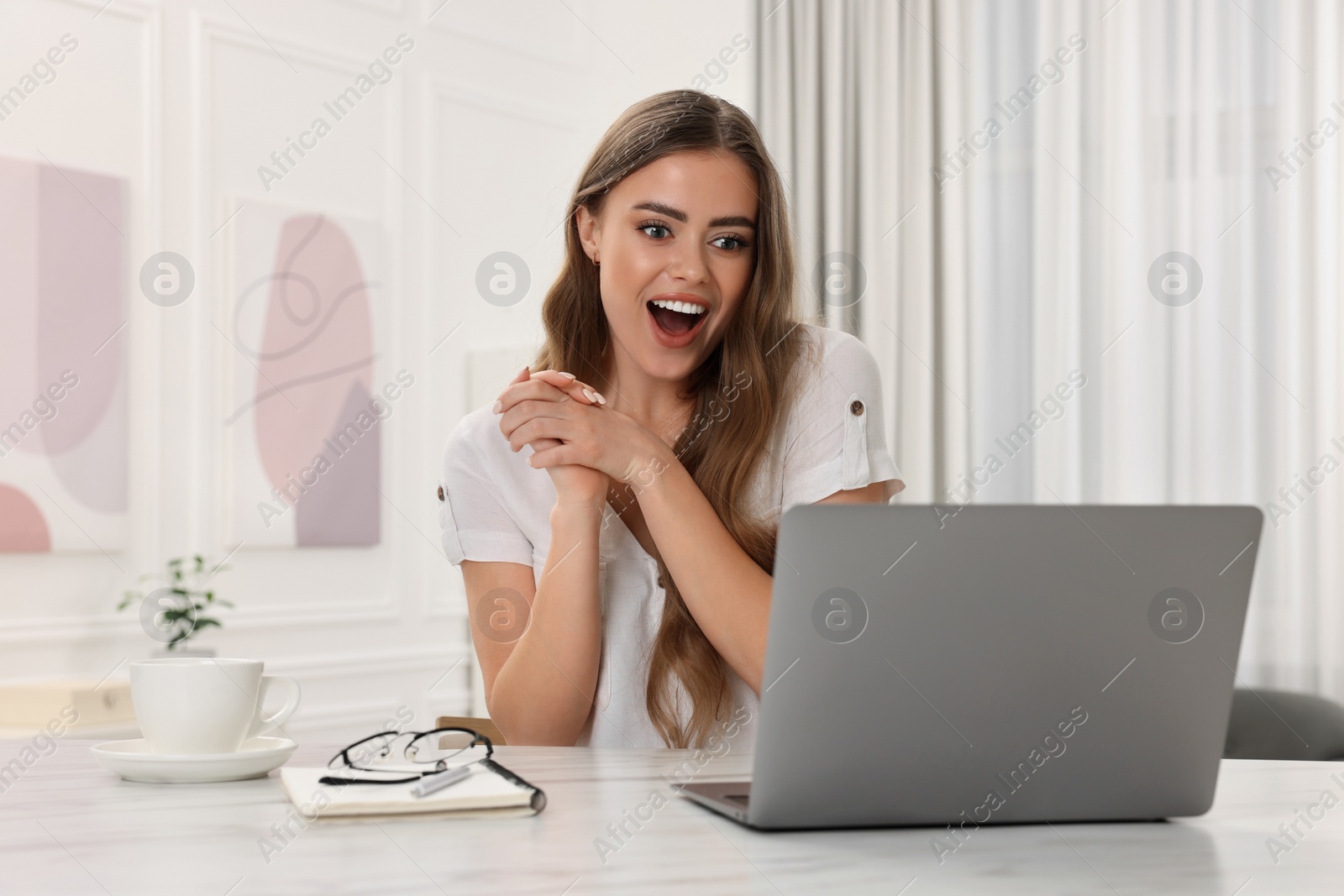 Photo of Surprised woman using laptop at white table in room