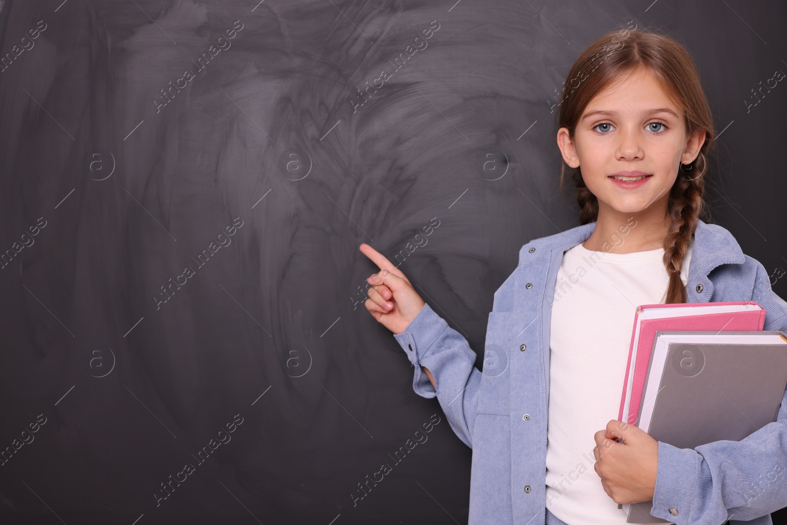 Photo of Smiling schoolgirl with books pointing at something on blackboard. Space for text