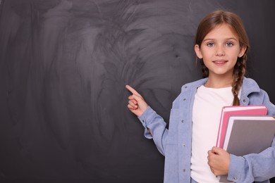 Photo of Smiling schoolgirl with books pointing at something on blackboard. Space for text