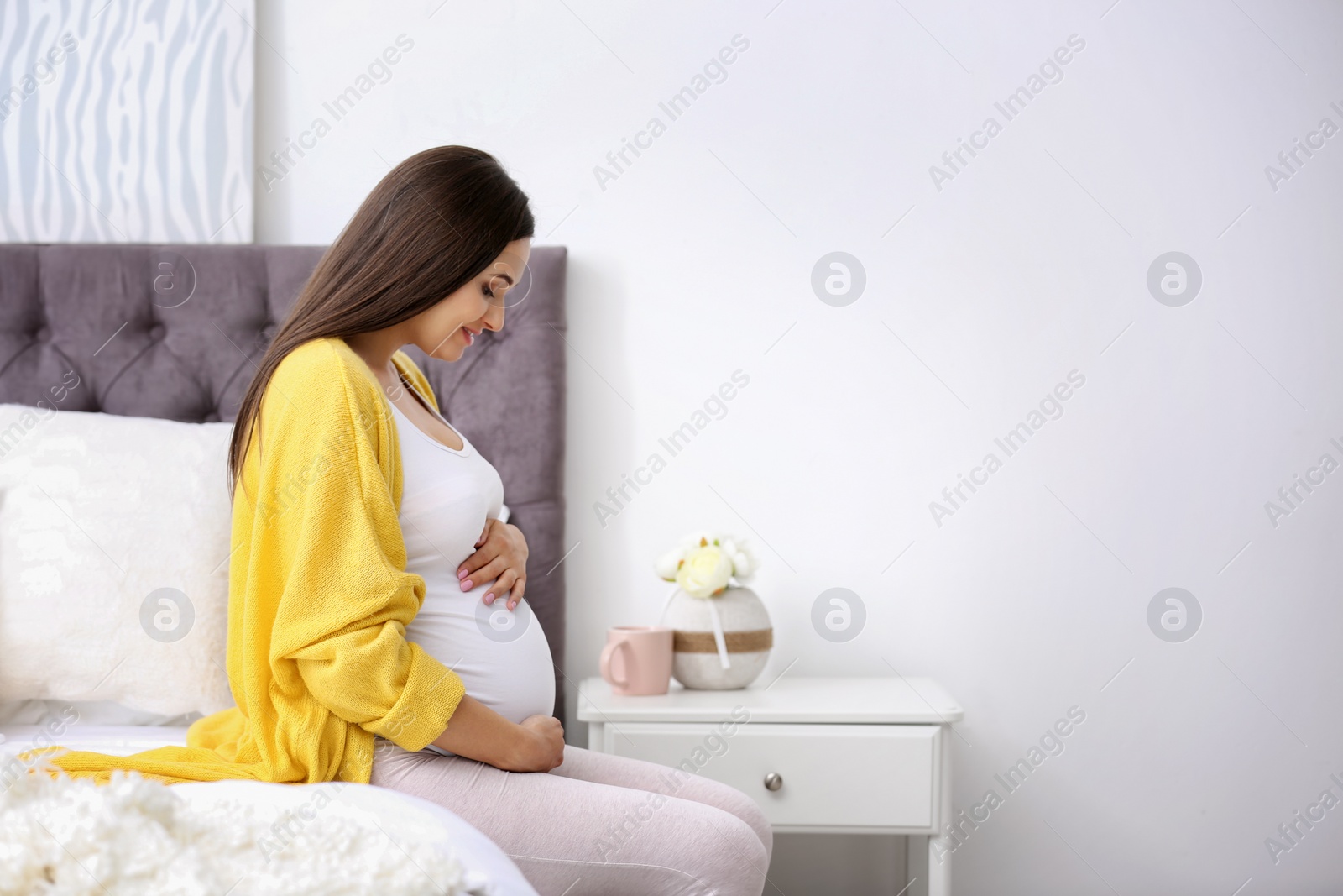 Photo of Young beautiful pregnant woman sitting on bed and touching her belly at home