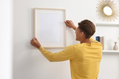 Young man hanging picture frame on white wall indoors, back view