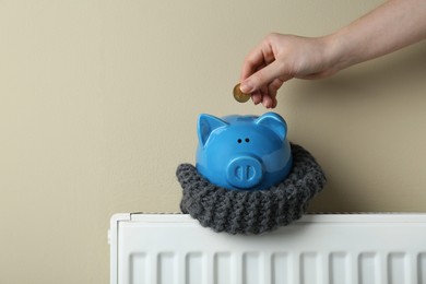 Photo of Woman putting coin into piggy bank on heating radiator against beige background, closeup. Space for text