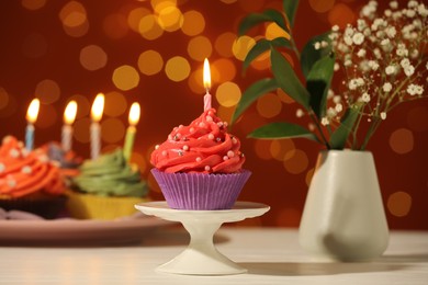 Photo of Stand with birthday cupcake on white table against blurred lights