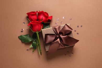 Photo of Beautiful gift box, flowers and confetti on brown background, flat lay