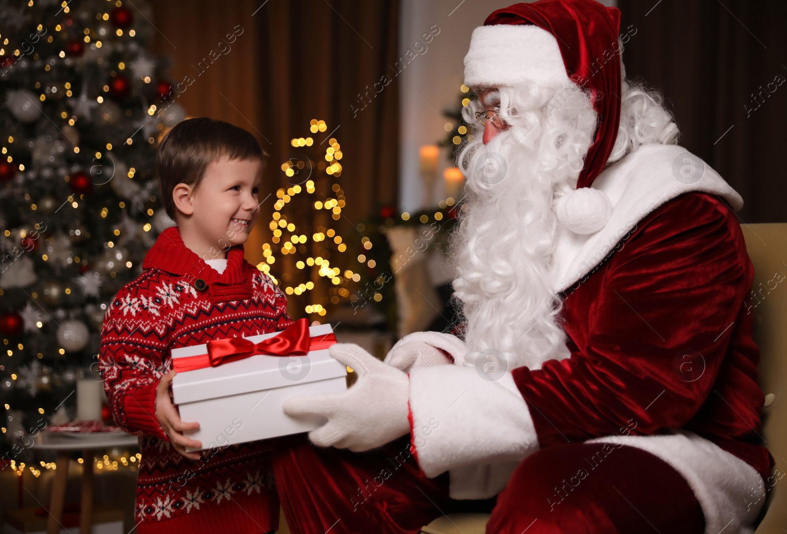 Photo of Santa Claus giving present to little boy in room decorated for Christmas