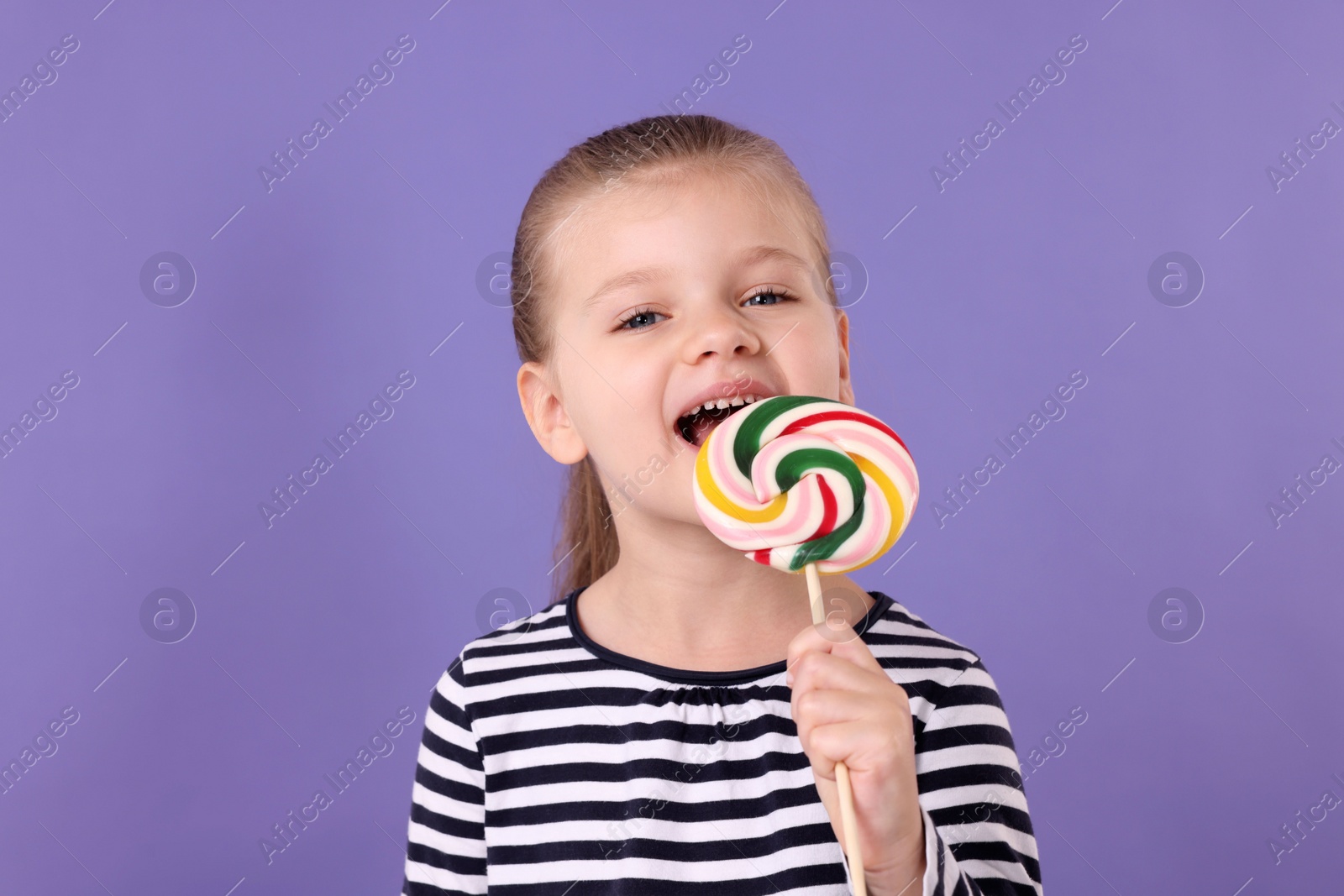 Photo of Happy little girl licking colorful lollipop swirl on violet background