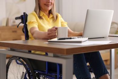 Photo of Woman in wheelchair with cup of drink using laptop at home, closeup