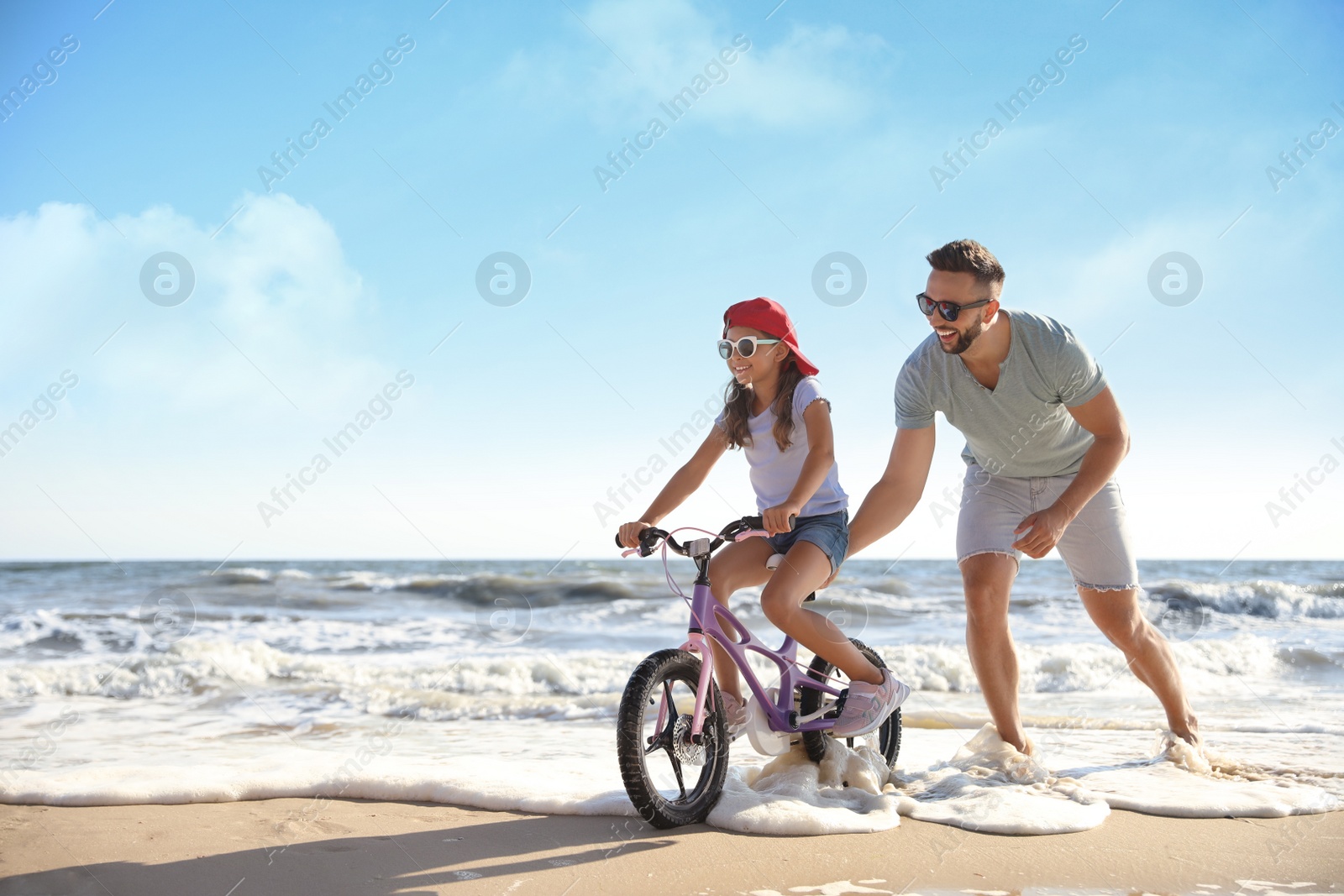 Photo of Happy father teaching daughter to ride bicycle on sandy beach near sea