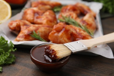 Photo of Fresh marinade, basting brush and raw marinated chicken wings on wooden table, closeup