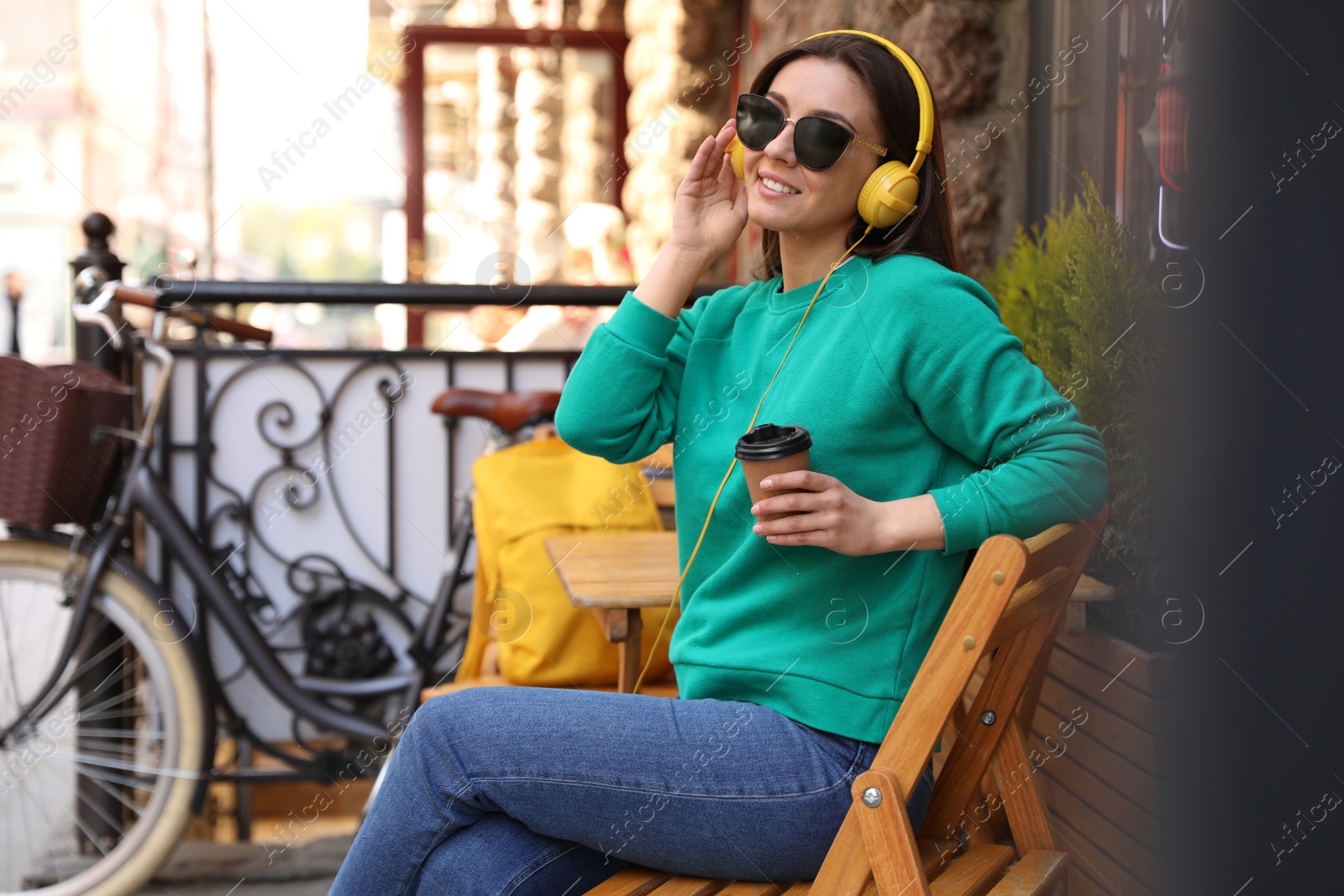 Photo of Happy young woman with coffee and headphones listening to music in outdoor cafe