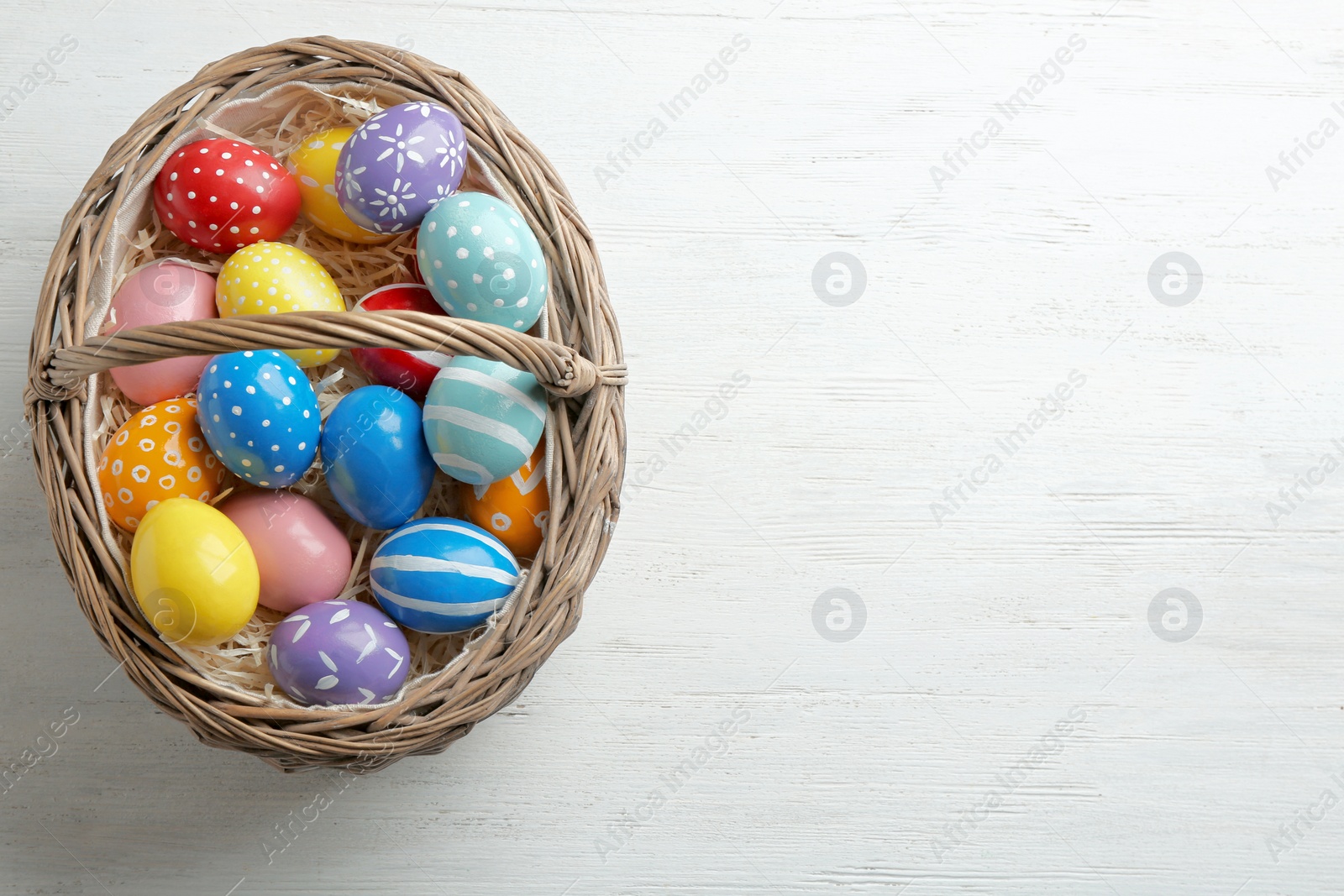 Photo of Wicker basket with painted Easter eggs on wooden table, top view. Space for text