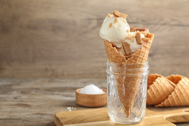 Photo of Waffle cone with ice cream, caramel and nuts in glass jar on wooden table. Space for text