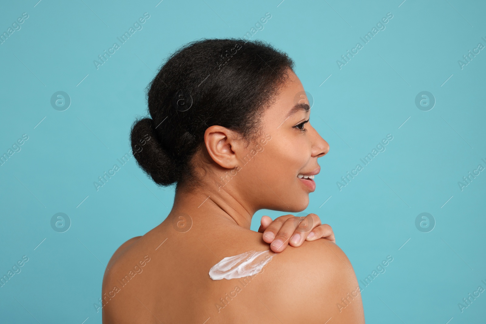 Photo of Young woman applying body cream onto back on light blue background