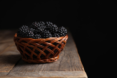 Photo of Fresh ripe blackberries in wicker bowl on wooden table against dark background. Space for text
