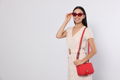 Photo of Beautiful young woman with red stylish bag in sunglasses on light grey background, space for text