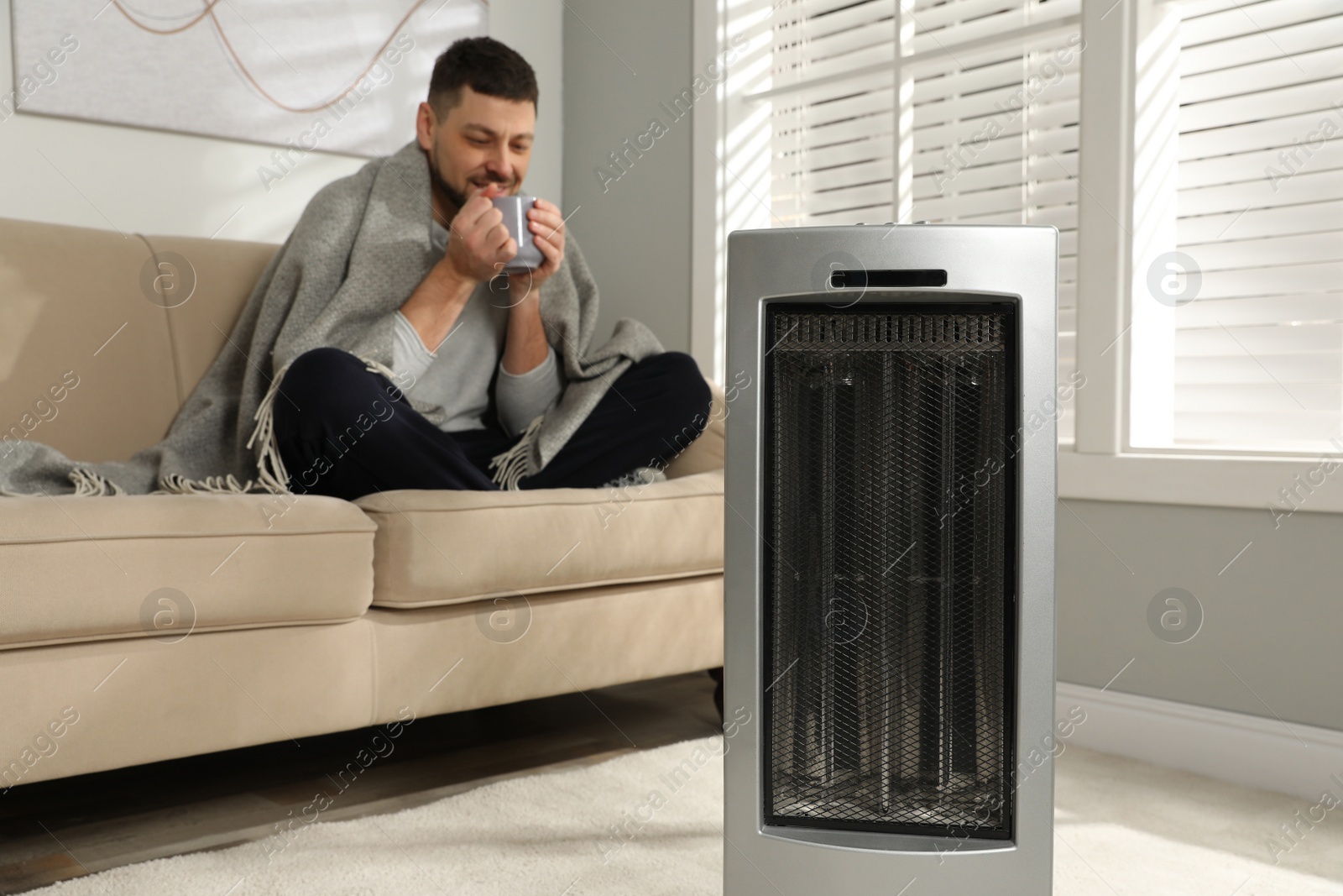 Photo of Man with cup of hot drink in living room, focus on electric ultrared heater