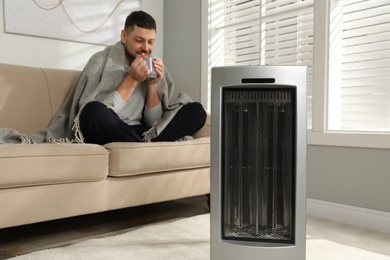 Man with cup of hot drink in living room, focus on electric ultrared heater