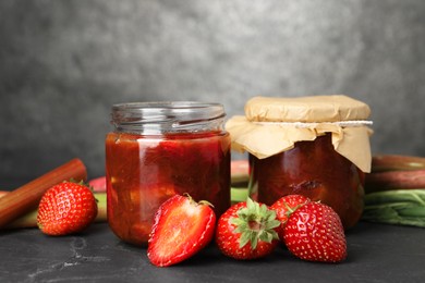 Jars of tasty rhubarb jam, fresh stems and strawberries on dark textured table