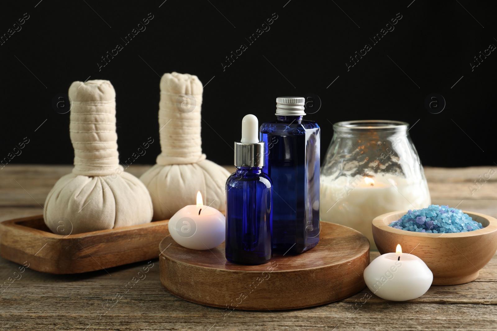 Photo of Spa composition. Cosmetic products, herbal bags, burning candles and sea salt on wooden table against black background