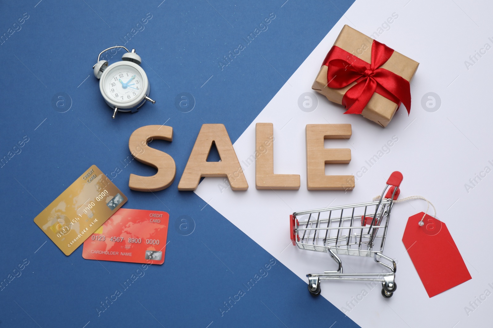 Photo of Word Sale made of wooden letters, shopping cart with tag, gift box, alarm clock and credit cards on color background, flat lay