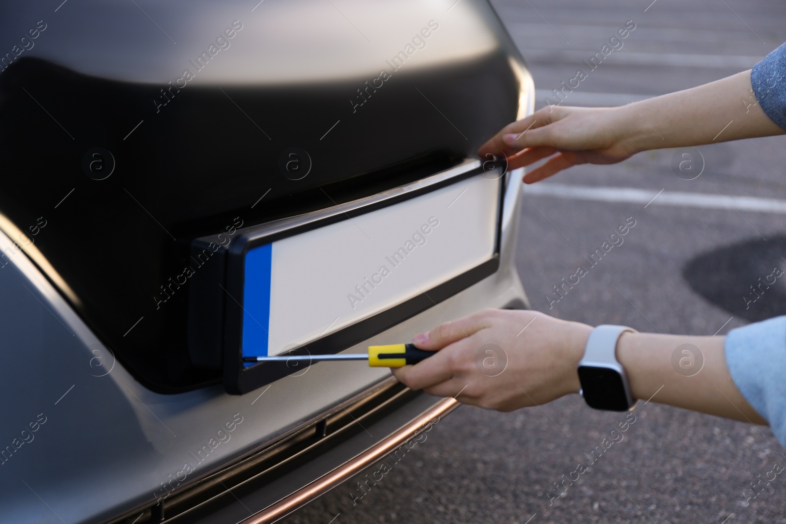 Photo of Woman with screwdriver installing vehicle registration plate to car outdoors, closeup