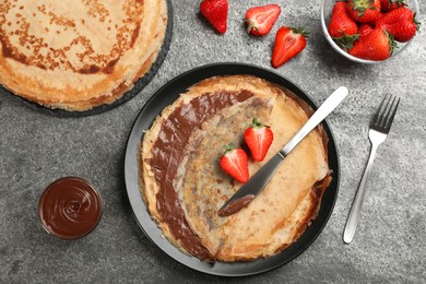 Photo of Delicious thin pancakes served with chocolate paste and strawberries on grey table, flat lay