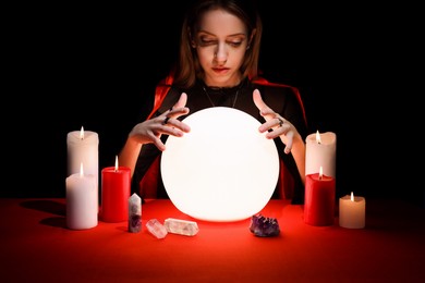 Photo of Soothsayer using glowing crystal ball to predict future at table in darkness. Fortune telling