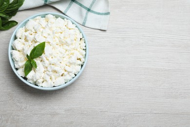 Fresh cottage cheese with basil in bowl on light wooden table, flat lay. Space for text