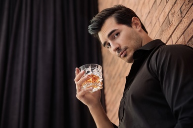 Young man with glass of whiskey near brick wall indoors. Space for text
