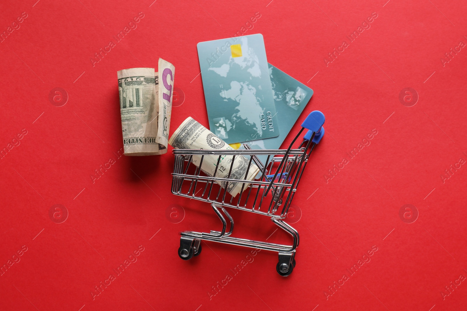 Photo of Small metal shopping cart with credit cards and dollar banknotes on red background, flat lay