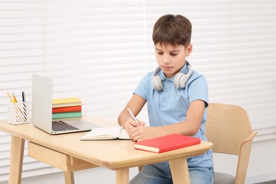 Boy writing in notepad near laptop at desk in room. Home workplace