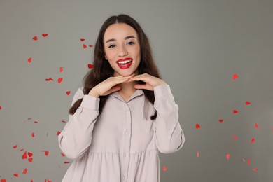 Beautiful young woman under falling heart shaped confetti on grey background