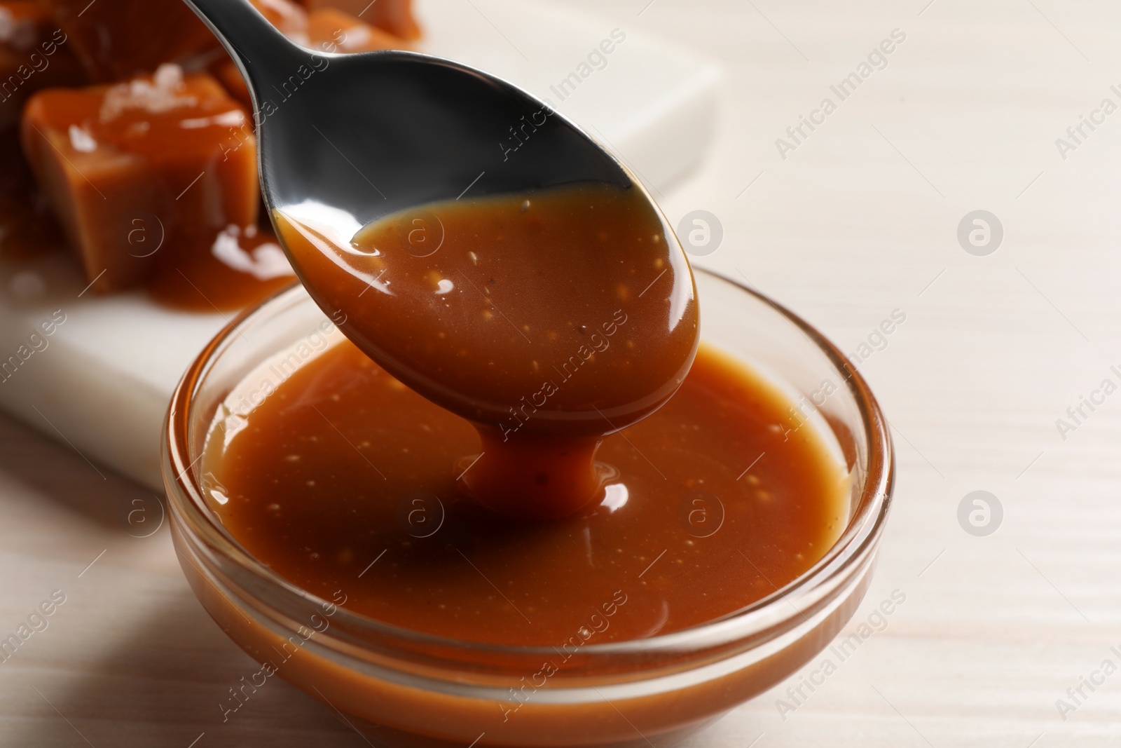 Photo of Taking tasty salted caramel with spoon from glass bowl at white wooden table, closeup