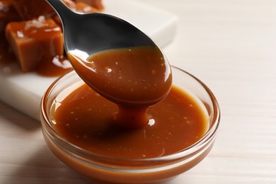 Photo of Taking tasty salted caramel with spoon from glass bowl at white wooden table, closeup