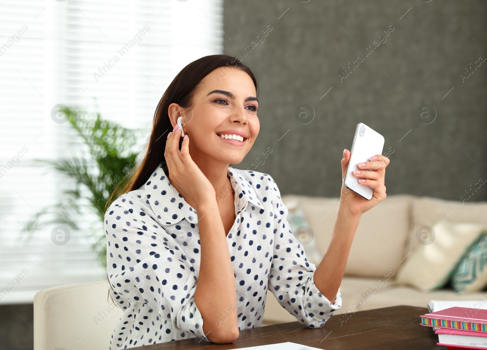 Photo of Happy young woman with smartphone listening to music through wireless earphones at table in office