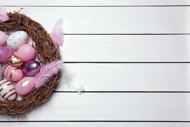 Festively decorated Easter eggs, vine wreath and feathers on white wooden table, top view. Space for text