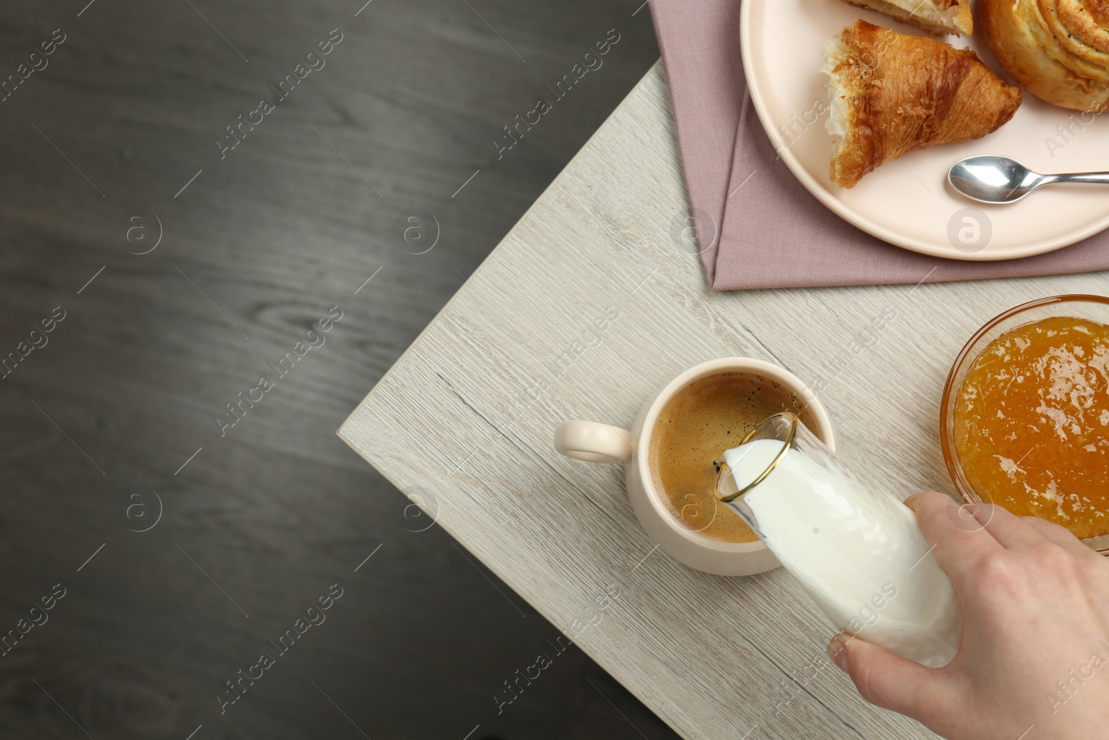 Photo of Woman pouring milk into cup with coffee and pastry at wooden table, top view. space for text