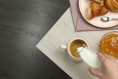 Woman pouring milk into cup with coffee and pastry at wooden table, top view. space for text