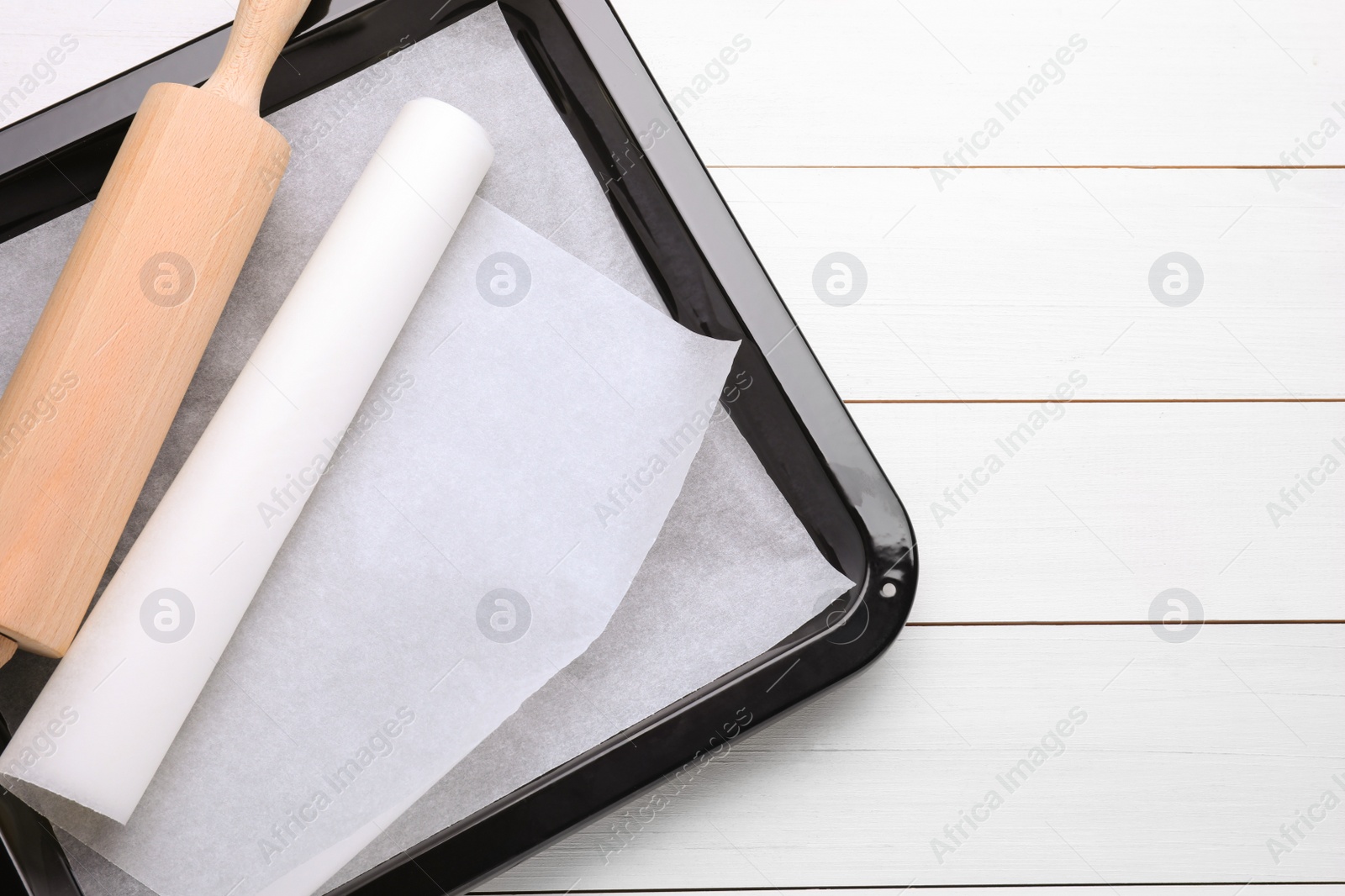 Photo of Baking pan with parchment paper and rolling pin on white wooden table, top view. Space for text