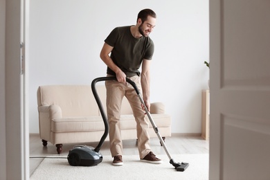 Photo of Young man cleaning carpet with vacuum in living room