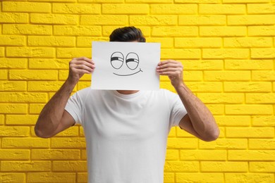 Photo of Man hiding emotions using card with drawn smiling face near yellow brick wall