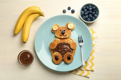Photo of Creative serving for kids. Plate with cute bear made of pancakes, blueberries, bananas and chocolate paste on light wooden table, flat lay