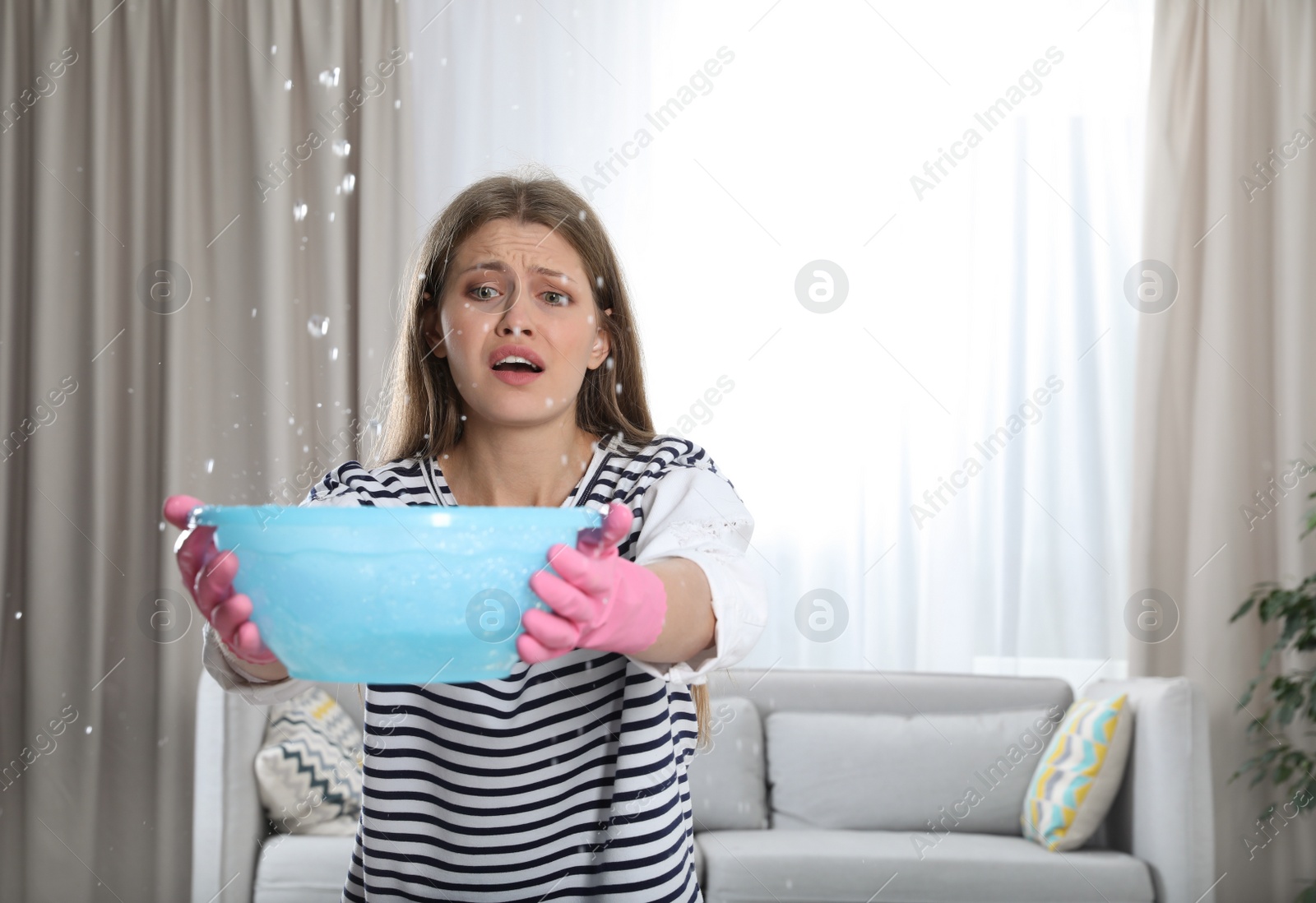 Photo of Emotional young woman collecting water leaking from ceiling in living room. Plumber service