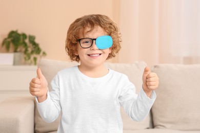 Photo of Happy boy with eye patch on glasses showing thumbs up indoors. Strabismus treatment