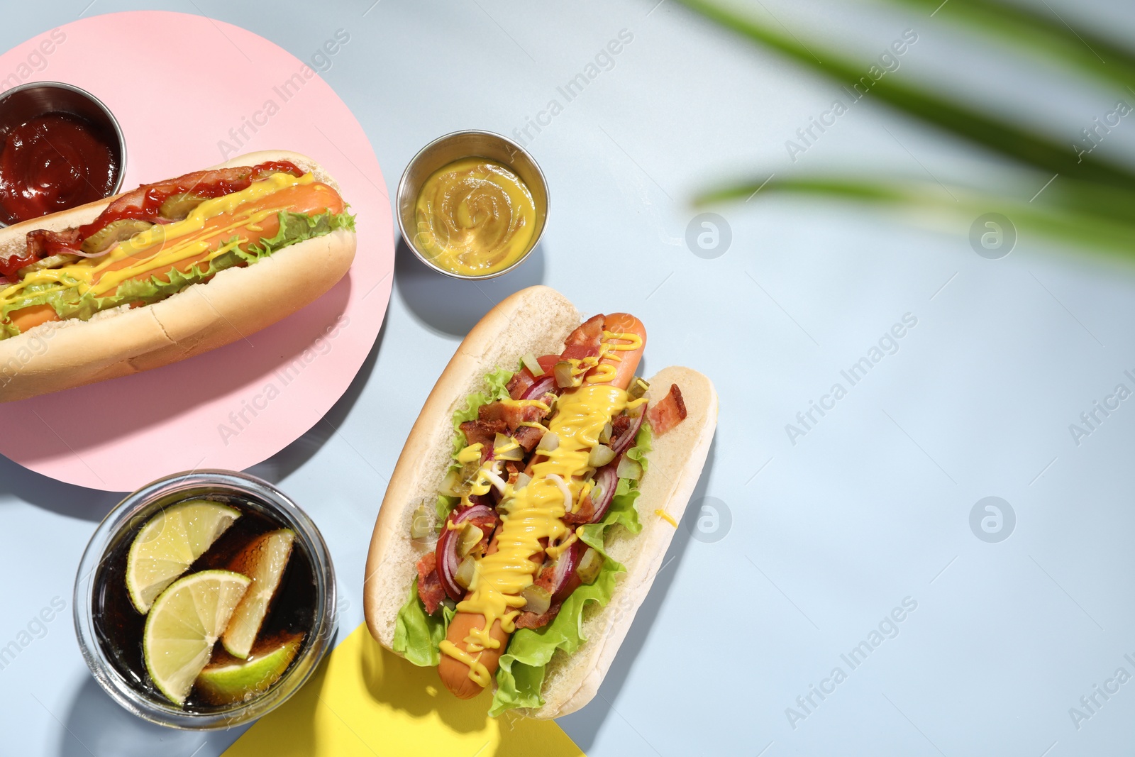 Photo of Delicious hot dogs with bacon, lettuce, onion and pickle served on light blue table, flat lay. Space for text