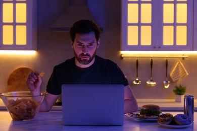 Photo of Man eating chips while using laptop in kitchen at night. Bad habit