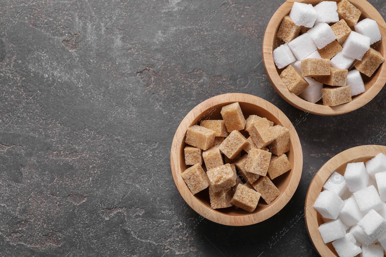 Photo of Different sugar cubes in bowls on gray textured table, flat lay. Space for text